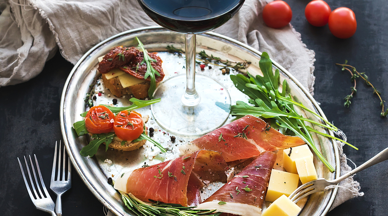 Wine appetizer set. Glass of red wine, vintage dinnerware, brushetta with cherry, dried tomatoes, arugula, parmesan, smoked meat on silver tray over rustic grunge surface. Closeup